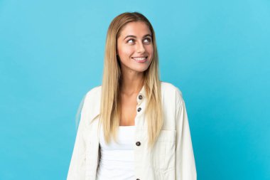 Mavi arka planda izole edilmiş genç sarışın kadın yukarı bakarken bir fikir düşünüyor.