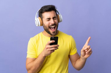 Mor arka planda sakallı genç yakışıklı bir adam cep telefonuyla müzik dinliyor ve şarkı söylüyor.