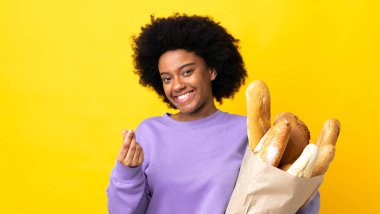 Genç Afrikalı Amerikalı bir kadın sarı arka planda izole edilmiş bir ekmek alıyor ve para hareketi yapıyor.