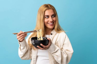 Genç Uruguaylı sarışın kadın izole edilmiş mavi arka planda elinde çubuklu bir kase erişteyle yukarı bakıyor.