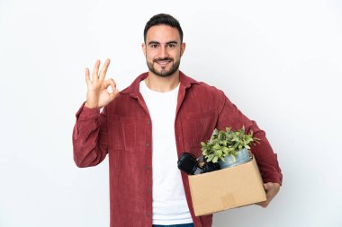 Young caucasian man making a move while picking up a box full of things isolated on white background showing ok sign with fingers