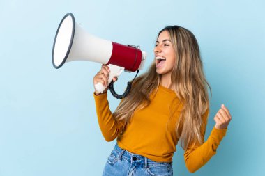 İzole edilmiş mavi arka planda genç İspanyol kadın megafondan bir şey duyurmak için bağırıyor.