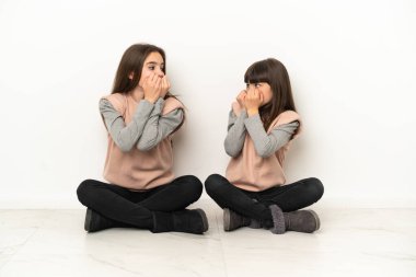 Beyaz arka planda izole bir şekilde yerde oturan küçük kız kardeşler biraz gergin ve korkmuş durumda.