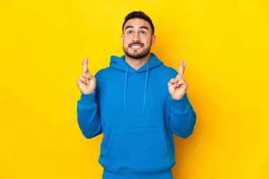 Sarı arka planda izole edilmiş beyaz, yakışıklı genç bir adam parmaklarını çapraz yapıp en iyisini diliyor.