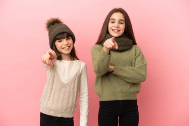 Pembe arka planda izole edilmiş kışlık giysiler giyen küçük kız kardeşler kendilerine güvenen bir ifadeyle parmaklarını sana doğrultuyorlar.