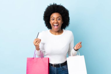 Genç Afrikalı Amerikalı bir kadın mavi arka planda izole edilmiş elinde alışveriş çantaları ve şaşırmış