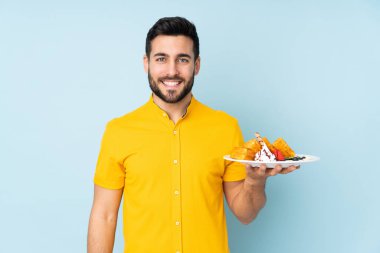 Mavi arka planda izole edilmiş waffle 'lar tutan beyaz adam çok gülümsüyor.
