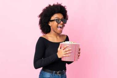 Pembe arka planda 3 boyutlu gözlüklü ve elinde koca bir kova patlamış mısır tutan genç Afrikalı Amerikalı kadın.