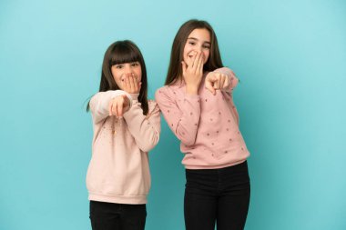 Küçük kız kardeşler mavi arka planda izole edilmiş. Birini işaret edip gülüyorlar.