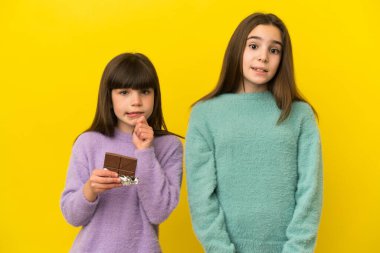 Sarı arka planda izole edilmiş küçük kız kardeşler çikolata tableti alıyorlar ve kuşkuları var.