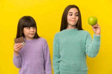 Sarı arka planda izole edilmiş küçük kız kardeşler bir elinde çikolata tableti, diğer elinde elma tutarken şüpheye düşüyorlar.
