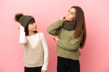 Pembe arka planda izole edilmiş kışlık giysiler giyen küçük kız kardeşler kafa karıştırırken bir fikir üretirler.