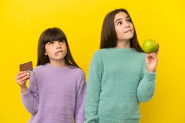 Sarı arka planda izole edilmiş küçük kız kardeşler bir elinde çikolata tableti, diğer elinde elma tutarken şüpheye düşüyorlar.
