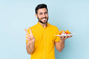 Mavi arka planda gözleme tutan beyaz adam gülümsüyor ve zafer işareti gösteriyor.