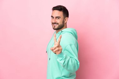 Beyaz tenli yakışıklı adam pembe arka planda izole edilmiş gülümsüyor ve zafer işareti gösteriyor.