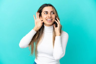 Mavi arka planda izole edilmiş cep telefonu kullanan beyaz bir kadın elini kulağına koyarak bir şeyler dinliyor.