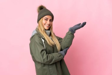 Pembe arka planda izole edilmiş, kış şapkalı genç bir kadın davet edilmek için ellerini yana uzatıyor.