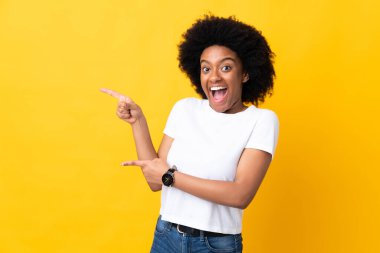Sarı arka planda izole edilmiş genç Afrikalı Amerikalı kadın şaşırmış ve işaret ediyor.