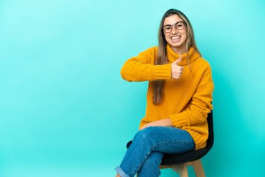 Mavi arka planda izole edilmiş bir sandalyede oturan beyaz bir kadın baş parmak işareti yapıyor.