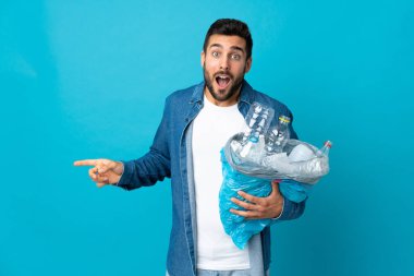 Mavi arka planda izole edilmiş plastik şişelerle dolu bir çanta tutan genç beyaz adam şaşırdı ve parmağını yan tarafa gösterdi.