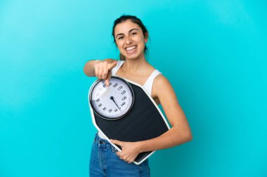 Mavi arka planda izole edilmiş beyaz bir kadın elinde bir tartı makinesiyle ön tarafı işaret ediyor.