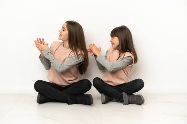 Beyaz arka planda izole bir şekilde yerde oturan küçük kız kardeşler bir konferanstan sonra alkışlıyorlar.