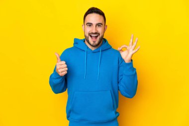 Sarı arka planda izole edilmiş beyaz, yakışıklı genç bir adam tamam işareti ve baş parmak hareketi gösteriyor.