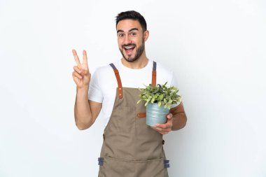 Bahçıvan elinde bir bitki tutuyor. Beyaz arka planda izole edilmiş. Gülümsüyor ve zafer işareti gösteriyor.