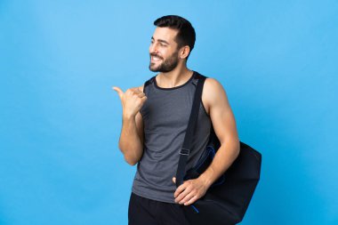 Spor çantalı genç adam mavi arka planda izole edilmiş bir ürün sunmak için kenarı işaret ediyor.