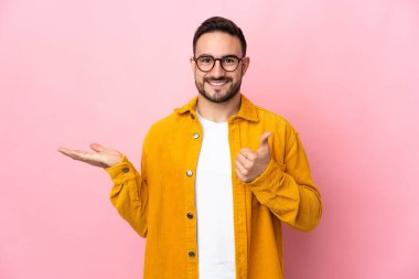 Young caucasian handsome man isolated on pink background holding copyspace imaginary on the palm to insert an ad and with thumbs up