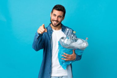 Mavi arka planda izole edilmiş plastik şişelerle dolu bir çanta taşıyan genç beyaz bir adam kendinden emin bir ifadeyle sana işaret ediyor.