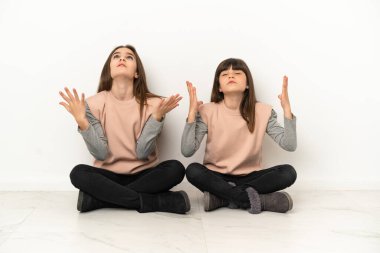 Beyaz arka planda izole bir şekilde yerde oturan küçük kız kardeşler kötü bir durum yüzünden hayal kırıklığına uğradılar.