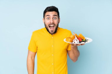 Mavi arka planda, şaşırmış ve şok olmuş bir yüz ifadesiyle waffle tutan beyaz bir adam.