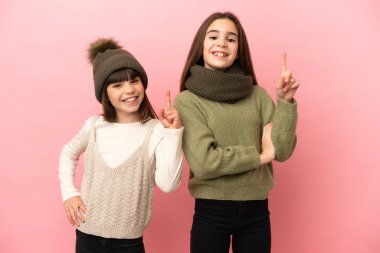 Pembe arka planda izole edilmiş kışlık giysiler giyen küçük kız kardeşler en iyinin işareti olarak parmağını kaldırıyor.