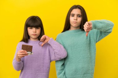 Sarı arka planda izole edilmiş küçük kız kardeşler çikolata tableti alarak kötü sinyal veriyorlar.