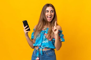 İzole edilmiş sarı arka plan üzerinde cep telefonu kullanan genç bir kadın parmak kaldırırken çözümü bulmayı umuyor.