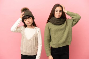 Pembe arka planda izole edilmiş kışlık giysiler giyen küçük kız kardeşler hayal kırıklığı ifadesi gösteriyorlar ve anlamıyorlar.