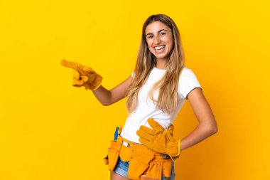 Genç İspanyol elektrikçi kadın, izole edilmiş sarı duvarın üzerinden parmak göstererek bir ürün sunuyor.