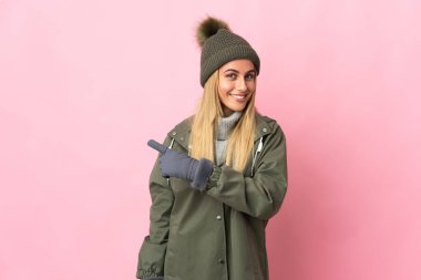 Pembe arka planda izole edilmiş, kış şapkalı genç bir kadın bir ürün sunmak için kenarı işaret ediyor.