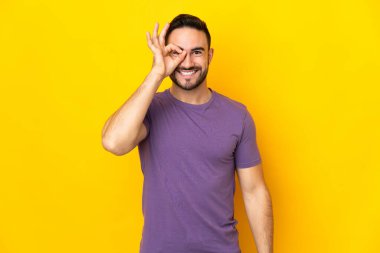 Sarı arka planda izole edilmiş beyaz, yakışıklı genç bir adam parmaklarıyla tamam işareti gösteriyor.