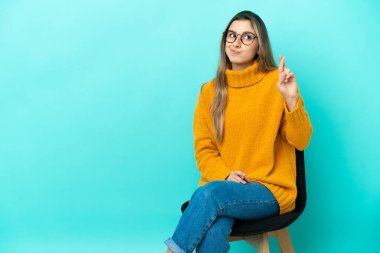 Mavi arka planda izole edilmiş bir sandalyede oturan beyaz bir kadın, şans diliyor.