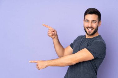 Beyaz tenli yakışıklı adam, bir tarafa işaret ediyor ve izole edilmiş mor arka planda bir ürün sunuyor.