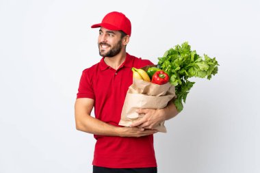 Sakallı genç adam elinde sebze dolu bir çanta beyaz arka planda izole edilmiş bir şekilde yan tarafa bakıyor.