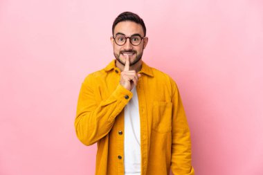 Pembe arka planda izole edilmiş beyaz, yakışıklı genç bir adam sessiz bir jest işareti gösteriyor. Parmağını ağzına sokuyor.