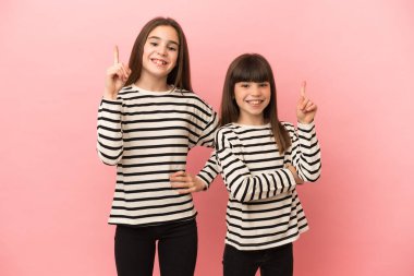 Pembe arka planda izole edilmiş küçük kız kardeşler en iyinin işareti olarak parmağını kaldırıyor.