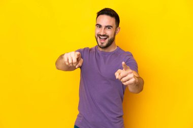 Sarı arka planda izole edilmiş beyaz, yakışıklı genç adam mutlu bir ifadeyle ön tarafı işaret ediyor.