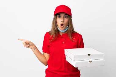 Pizza teslimatı yapan kadın elinde pizza tutuyordu. Koronavirüsten koruyordu. İzole edilmiş beyaz duvarın üzerinde bir maske vardı.