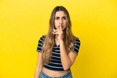 Sarı arka planda izole edilmiş genç beyaz bir kadın sessiz bir jest işareti gösteriyor. Parmağını ağzına sokuyor.