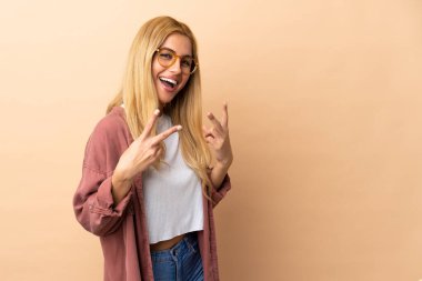 Genç Uruguaylı sarışın kadın izole edilmiş arka planda gülümsüyor ve zafer işareti gösteriyor.