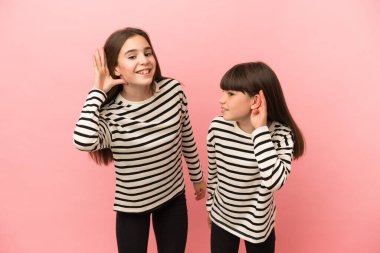 Küçük kız kardeşler pembe arka planda izole edilmiş bir şekilde ellerini kulağına koyarak bir şeyler dinliyorlar.